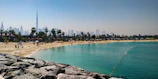 A sandy beach with sunbathers and palm trees, adjacent to a calm turquoise sea. In the distance, a modern city skyline with skyscrapers, including a notably tall building. The foreground features rocky beachfront barriers.