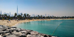 A sandy beach with sunbathers and palm trees, adjacent to a calm turquoise sea. In the distance, a modern city skyline with skyscrapers, including a notably tall building. The foreground features rocky beachfront barriers.