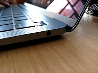A close-up view of a laptop keyboard with a focus on the side edge, highlighting the sleek design and metallic finish. The laptop is placed on a wooden surface, and the screen is slightly raised, showing reflections.