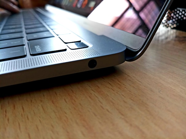 A close-up view of a laptop keyboard with a focus on the side edge, highlighting the sleek design and metallic finish. The laptop is placed on a wooden surface, and the screen is slightly raised, showing reflections.