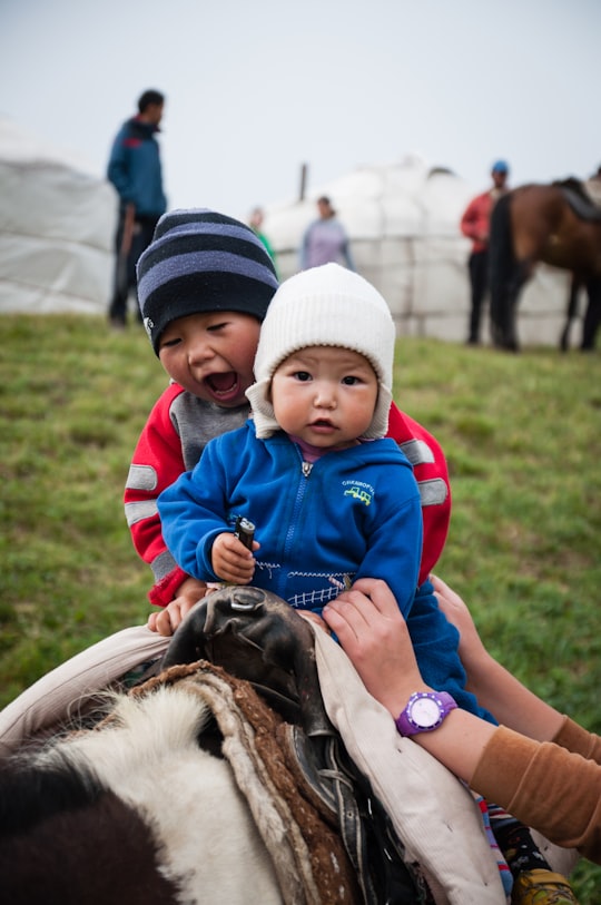 boy in blue jacket and white hat sitting on white and black horse during daytime in Kochkor Kyrgyzstan