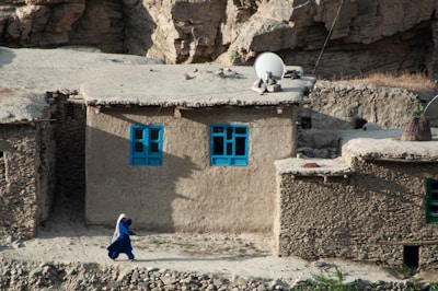 A rural clay or stone house with bright blue window frames is nestled against a rocky landscape. A woman dressed in a blue garment walks along a dirt path in front of the house. A satellite dish is mounted on the roof, and there are baskets and other household items nearby.