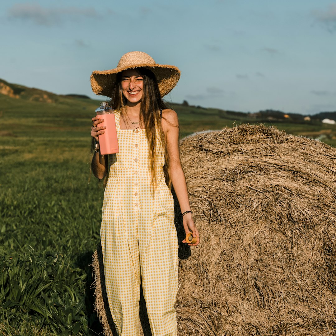 woman in brown and white plaid dress wearing brown straw hat standing on brown grass field