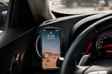 A smartphone is mounted on a car dashboard, displaying the time 8:30. The phone's wallpaper features a desert landscape with rocky terrain under a clear sky. The steering wheel and part of the car's instrument panel, including a speedometer and other gauges, are visible.