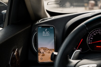 A smartphone is mounted on a car dashboard, displaying the time 8:30. The phone's wallpaper features a desert landscape with rocky terrain under a clear sky. The steering wheel and part of the car's instrument panel, including a speedometer and other gauges, are visible.
