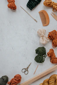 A collection of crafting tools and materials on a textured surface, including yarn in various colors, a wooden comb, metal scissors, knitting needles, and a ruler.