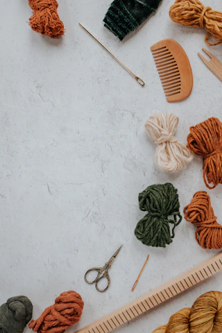 A collection of crafting tools and materials on a textured surface, including yarn in various colors, a wooden comb, metal scissors, knitting needles, and a ruler.
