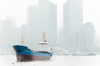 A cargo ship with a blue and red hull sails along a river, framed against the backdrop of a city skyline filled with tall, modern skyscrapers shrouded in mist or fog. The scene appears calm with the water reflecting the overcast sky.