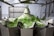 A metallic machine is processing green leafy vegetables, likely in an industrial setting. The setup includes several compartments or trays arranged in a circular pattern, with multiple pieces of lettuce or similar greens on top. Stainless steel components are visible, suggesting a food processing environment.