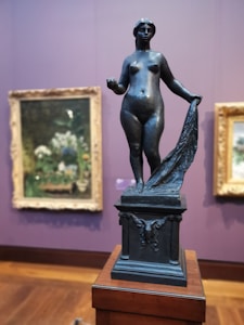 A bronze sculpture of a nude female figure stands atop a detailed plinth, holding a draped cloth in one hand. The background features blurred framed paintings with floral motifs on a purple wall. The setting suggests an art gallery or museum.