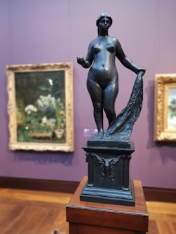 A bronze sculpture of a nude female figure stands atop a detailed plinth, holding a draped cloth in one hand. The background features blurred framed paintings with floral motifs on a purple wall. The setting suggests an art gallery or museum.