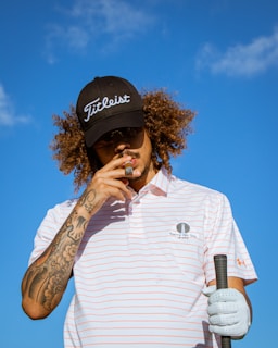 A person with curly hair wearing a black cap and sunglasses is holding a cigar in one hand and a golf club in the other. The person wears a striped polo shirt with visible tattoos on one arm. The background is a clear blue sky.