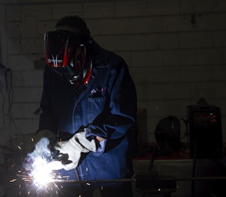 A person wearing protective gear, including a helmet and gloves, is engaged in welding. Bright sparks and a white light emanate from the welding point. The background is dimly lit, revealing a workshop environment with tools and equipment visible.