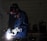 A person wearing protective gear, including a helmet and gloves, is engaged in welding. Bright sparks and a white light emanate from the welding point. The background is dimly lit, revealing a workshop environment with tools and equipment visible.