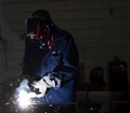 A person wearing protective gear, including a helmet and gloves, is engaged in welding. Bright sparks and a white light emanate from the welding point. The background is dimly lit, revealing a workshop environment with tools and equipment visible.