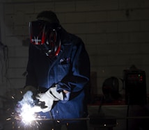 A person wearing protective gear, including a helmet and gloves, is engaged in welding. Bright sparks and a white light emanate from the welding point. The background is dimly lit, revealing a workshop environment with tools and equipment visible.