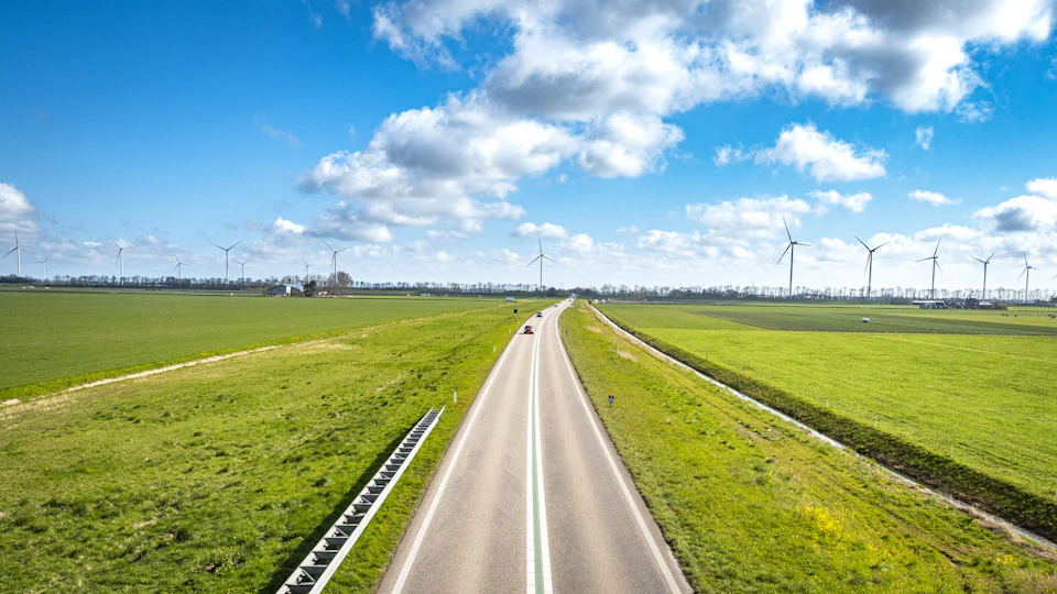 Serene rural landscape with two-lane road flanked by lush green fields and wind turbines