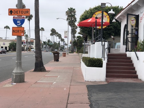 A street scene with palm trees lining the road. There are traffic signs indicating a detour on Interstate 5 North. A restaurant with a red umbrella sits on the right side, and a trash bin is visible on the sidewalk.