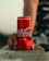 A close-up view of a hand holding a red can of Coca-Cola resting on a concrete surface. The background is blurred, drawing attention to the vibrant color and branding of the can.