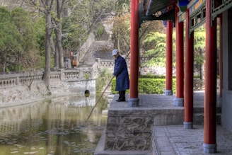 A person stands on a stone platform next to a body of water, using a long stick to clean the water surface. They are dressed in a blue coat and cap. The setting includes traditional architectural elements with red pillars and intricate stone carvings. Lush greenery and stone railings line the background.