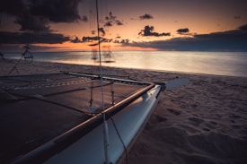 A tranquil seascape at sunset with a catamaran resting on the sandy beach. The sky transitions from deep blue to orange and pink hues as the sun sets on the horizon. Gentle waves lap the shore, and dark clouds are scattered in the sky.