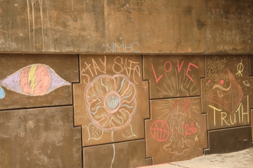 A series of chalk drawings and words on a concrete wall. On the left, there is a large eye with a lightning bolt through it. Next to it is a flower-like design with 'STAY SAFE' written around it. In the center, the word 'LOVE' is prominently displayed with radiating lines. To the right, there's a drawing of a candle with a red basketball and the number '26' underneath. The word 'TRUTH' is written in bold letters on the far right, accompanied by abstract symbols and shapes.