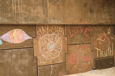 A series of chalk drawings and words on a concrete wall. On the left, there is a large eye with a lightning bolt through it. Next to it is a flower-like design with 'STAY SAFE' written around it. In the center, the word 'LOVE' is prominently displayed with radiating lines. To the right, there's a drawing of a candle with a red basketball and the number '26' underneath. The word 'TRUTH' is written in bold letters on the far right, accompanied by abstract symbols and shapes.