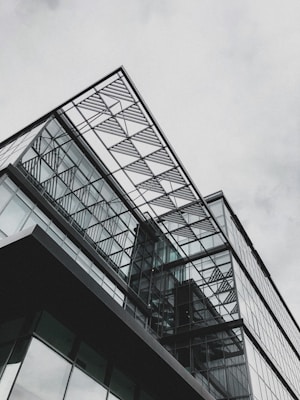 A modern glass building featuring geometric patterns and sharp angles. The architectural design includes large glass panels and metal frames, creating a sleek and contemporary appearance against a gray sky.