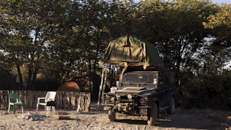 A rugged off-road camper trailer parked by a forested lake at sunrise.