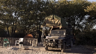 A rugged off-road vehicle with a rooftop tent is parked on sandy ground, surrounded by trees. The campsite includes a rustic fence, a couple of plastic chairs, a small barbecue grill, and camping tents. The light suggests it's either early morning or late afternoon, creating a serene atmosphere.