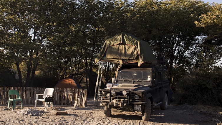 A rugged off-road vehicle with a rooftop tent is parked on sandy ground, surrounded by trees. The campsite includes a rustic fence, a couple of plastic chairs, a small barbecue grill, and camping tents. The light suggests it's either early morning or late afternoon, creating a serene atmosphere.