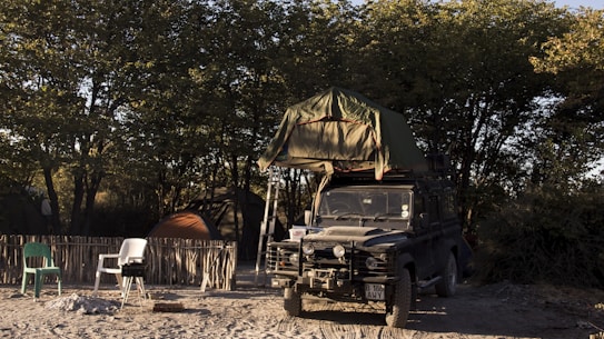 A rugged off-road vehicle with a rooftop tent is parked on sandy ground, surrounded by trees. The campsite includes a rustic fence, a couple of plastic chairs, a small barbecue grill, and camping tents. The light suggests it's either early morning or late afternoon, creating a serene atmosphere.