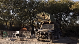 A rugged off-road vehicle with a rooftop tent is parked on sandy ground, surrounded by trees. The campsite includes a rustic fence, a couple of plastic chairs, a small barbecue grill, and camping tents. The light suggests it's either early morning or late afternoon, creating a serene atmosphere.