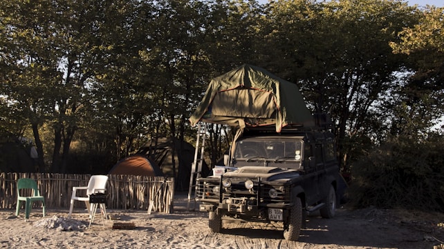 A rugged off-road vehicle with a rooftop tent is parked on sandy ground, surrounded by trees. The campsite includes a rustic fence, a couple of plastic chairs, a small barbecue grill, and camping tents. The light suggests it's either early morning or late afternoon, creating a serene atmosphere.
