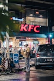 A street vendor stands beside a blue cart with the sign 'OTAK OTAK 2', and a nearby parked Honda car is visible. The scene is set in front of a KFC restaurant in an urban environment during nighttime. The atmosphere is bustling with a mix of modern commercial elements and local street commerce.