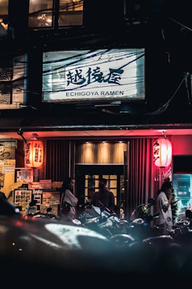 An outdoor night scene of a ramen restaurant with a sign that reads 'Echigoya Ramen' in prominent bold letters above. Red lanterns flank the entrance, illuminating the area with a warm glow. People are gathered outside, possibly waiting or chatting, while motorcycles are parked in the foreground. The ambiance suggests a busy and vibrant street life atmosphere.