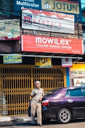A man holds a phone, leaning against a parked car in front of a store with multiple signs. The background features a variety of advertisements and signboards with vibrant colors.