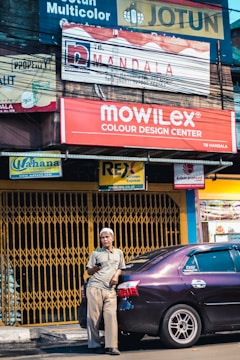 A man holds a phone, leaning against a parked car in front of a store with multiple signs. The background features a variety of advertisements and signboards with vibrant colors.