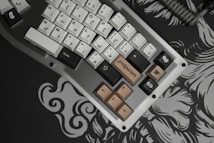 A mechanical keyboard with Arabic keycaps and a few novelty keycaps, including one labeled 'pr&eacute;parer,' is situated on an elaborately patterned desk mat. The keyboard features a combination of black, white, and brown keys, and the braided cable is visible as it attaches to the keyboard.