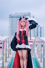 A person in a detailed cosplay outfit with pink hair and black cat ears stands confidently on an outdoor walkway. The costume features black with red accents, including ribbons and decorative elements, and is complemented by a white trim. This scene is set against a backdrop of modern urban buildings under a cloudy sky.