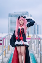 A person in a detailed cosplay outfit with pink hair and black cat ears stands confidently on an outdoor walkway. The costume features black with red accents, including ribbons and decorative elements, and is complemented by a white trim. This scene is set against a backdrop of modern urban buildings under a cloudy sky.