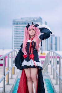A person in a detailed cosplay outfit with pink hair and black cat ears stands confidently on an outdoor walkway. The costume features black with red accents, including ribbons and decorative elements, and is complemented by a white trim. This scene is set against a backdrop of modern urban buildings under a cloudy sky.