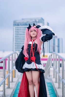 A person in a detailed cosplay outfit with pink hair and black cat ears stands confidently on an outdoor walkway. The costume features black with red accents, including ribbons and decorative elements, and is complemented by a white trim. This scene is set against a backdrop of modern urban buildings under a cloudy sky.