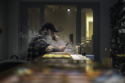 A person wearing a cap is sitting indoors at a desk, engrossed in their phone, while vaping. The atmosphere is dimly lit, with smoke creating a hazy effect around the individual. The background includes shelves stocked with various items and a partially open door revealing light.