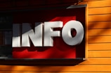 Large three-dimensional white letters spelling 'INFO' are mounted on a contrasting bright red background. The structure is attached to a wall with wooden planks painted in a vivid orange color, and gentle shadows are cast across the surface.
