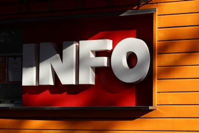 Large three-dimensional white letters spelling 'INFO' are mounted on a contrasting bright red background. The structure is attached to a wall with wooden planks painted in a vivid orange color, and gentle shadows are cast across the surface.