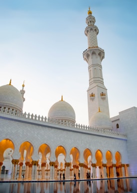 A grand mosque with stunning white domes and intricate arched designs. The tall minaret stands prominently against the pastel sky. Golden lighting highlights the architectural details, creating a serene and majestic ambiance.