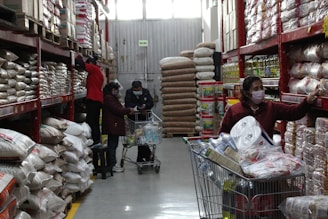 In a large warehouse-style store aisle, several people are shopping, pushing carts filled with various goods. Shelves are stocked with large bags of grains and boxes. People are wearing masks, indicative of health precautions.