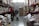 In a large warehouse-style store aisle, several people are shopping, pushing carts filled with various goods. Shelves are stocked with large bags of grains and boxes. People are wearing masks, indicative of health precautions.