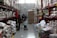 In a large warehouse-style store aisle, several people are shopping, pushing carts filled with various goods. Shelves are stocked with large bags of grains and boxes. People are wearing masks, indicative of health precautions.